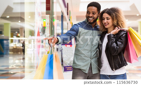 A joyful African American couple is engaged in a shopping spree, with the man pointing out something interesting to the woman, who is carrying multiple colorful shopping bags, empty space 115636730