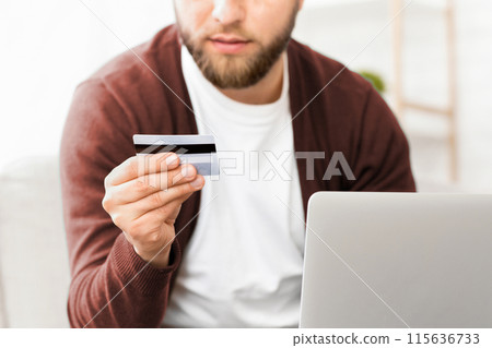 A man is shown holding a credit card in one hand and a laptop in the other. He appears to be in the process of making an online purchase or managing his finances, cropped A man is shown holding a credit card in one hand and a laptop in the other. He appears to be in the process of making an online purchase or managing his finances, cropped 115636733