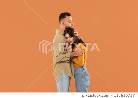 A young man and woman share an emotional embrace against a plain orange backdrop. Both appear thoughtful and comforted, capturing a moment of tenderness. 115636737