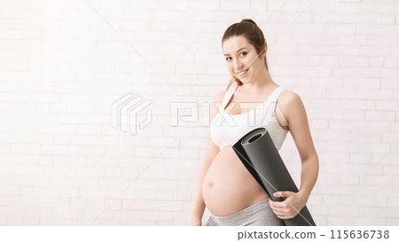 Sporty pregnant woman is standing while holding a yoga mat in her hands. She is visibly expecting and appears to be preparing for a yoga session, copy space 115636738