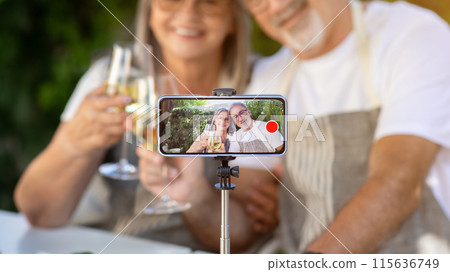 Smiling old caucasian husband and wife cheers glasses of wine in garden, shoot video on smartphone screen outdoor. Romantic date at home, dinner and relationship, holiday celebration with gadget 115636749