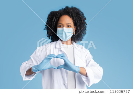 Glad young black woman doctor in white coat, protective mask and gloves shows heart sign with hand, isolated on blue studio background. Love, support, medicine, health care Glad young black woman doctor in white coat, protective mask and gloves shows heart sign with hand, isolated on blue studio background. Love, support, medicine, health care 115636757