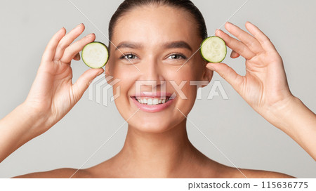 A young woman with a bright smile holds cucumber slices near her eyes against a plain background. She showcases a fresh and healthy skincare routine. A young woman with a bright smile holds cucumber slices near her eyes against a plain background. She showcases a fresh and healthy skincare routine. 115636775