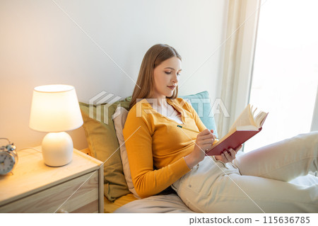 A girl is seated on a bed, engrossed in reading a book. She is dressed casually and appears focused on the content of the book. A girl is seated on a bed, engrossed in reading a book. She is dressed casually and appears focused on the content of the book. 115636785
