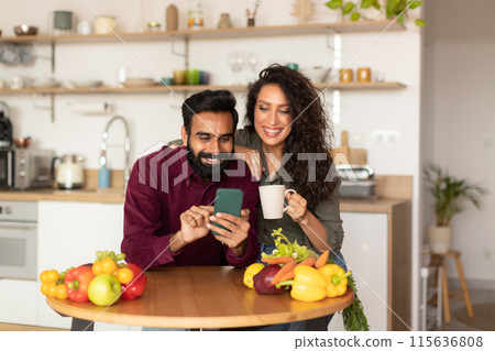Happy arab husband and wife relaxing in kitchen with smartphone and coffee, young couple using mobile phone, ordering food delivery or shopping online at home Happy arab husband and wife relaxing in kitchen with smartphone and coffee, young couple using mobile phone, ordering food delivery or shopping online at home 115636808