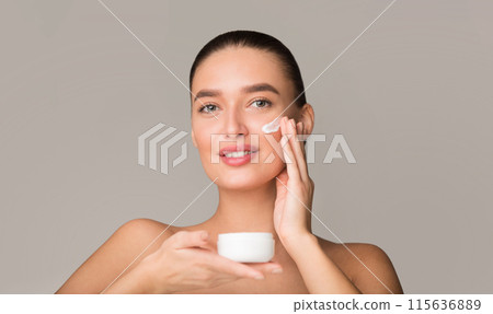 A woman is seen applying cream to her face in a well-lit room. She is using gentle, circular motions to massage the cream into her skin for hydration and nourishment. A woman is seen applying cream to her face in a well-lit room. She is using gentle, circular motions to massage the cream into her skin for hydration and nourishment. 115636889