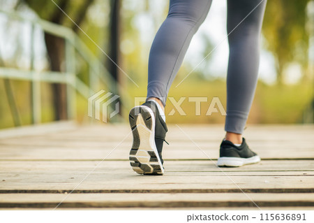 A person is taking a morning jog on a wooden pathway through a park. Wearing athletic gear, the runner steps confidently forward in black running shoes against a backdrop of green trees. 115636891