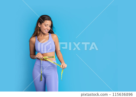 Positive millennial european lady in sportswear with measuring tape measures waist, isolated on blue studio background. Fit, sport, weight loss and diet result, ad and offer, lifestyle 115636896