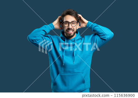 A man wearing a blue hoodie is standing with his hands pressed against his ears, appearing to be in distress or experiencing loud noise. He looks tense and focused on the sounds he is hearing. 115636900
