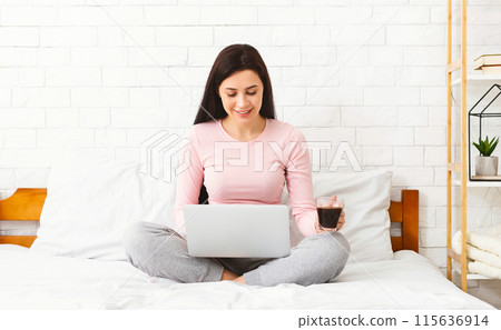 Brunette woman is seated on a bed, working on a laptop while holding a cup of coffee. She appears focused and engaged in her work, the laptop screen illuminating her face in the dimly lit room. Brunette woman is seated on a bed, working on a laptop while holding a cup of coffee. She appears focused and engaged in her work, the laptop screen illuminating her face in the dimly lit room. 115636914