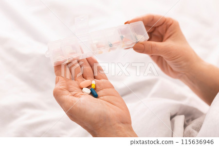 Cropped of woman holding a white pill bottle in one hand and a handful of assorted pills in the other hand, appears to be examining the pills closely, possibly preparing to take or organize them. Cropped of woman holding a white pill bottle in one hand and a handful of assorted pills in the other hand, appears to be examining the pills closely, possibly preparing to take or organize them. 115636946