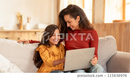 Joyful Middle Eastern Mom And Schoolgirl Daughter Using Laptop At Modern Living Room, Sitting With Computer, Browsing Internet And Having Fun Online At Home On Weekend. Panorama 115636976
