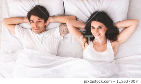 A young couple lies in bed together, seemingly after just waking up. They are both smiling and look relaxed and content. They are wearing casual clothes and are lying on white sheets and pillows. 115637007