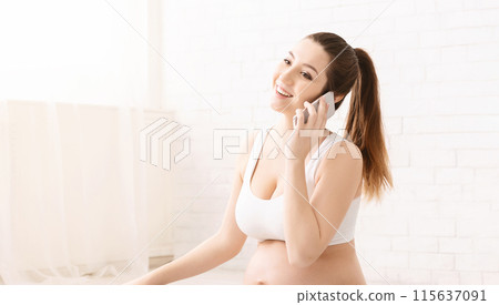 A pregnant woman is happily speaking on the phone while seated in a bright, airy room with white decor, wearing casual, light-colored clothing and appears to be in a positive mood, smiling widely. 115637091