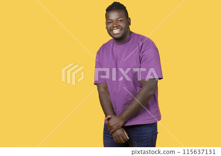 Shy African American young man looking at camera with toothy smile, holding hands together, posing wearing casual purple t-shirt, standing over yellow background, studio shot with free space 115637131