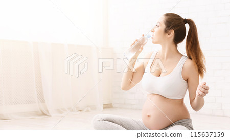A pregnant woman is sitting on the floor and drinking water while exercising. She holds a water bottle in her hand and appears to be staying hydrated, copy space 115637159