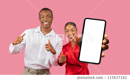 This image features a happy African American couple standing in front of a pink background. The woman is holding a smartphone with a blank screen in front of her, while the man is giving a thumbs-up 115637162