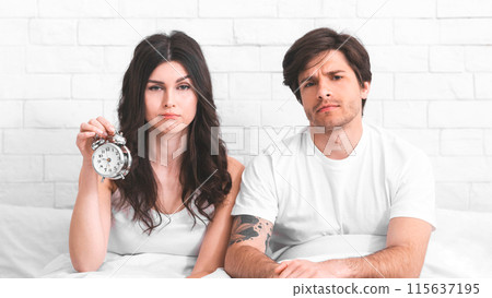 A young couple is sitting in bed, looking tired and unhappy after being woken up by an alarm clock. The woman is holding the alarm clock in her hand. They are both wearing white pajamas. 115637195