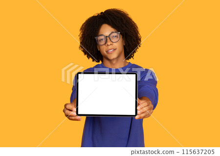 A young guy with curly hair and glasses wearing a blue sweatshirt is holding a blank tablet towards the camera against a solid yellow background. The focus is on the tablets screen. A young guy with curly hair and glasses wearing a blue sweatshirt is holding a blank tablet towards the camera against a solid yellow background. The focus is on the tablets screen. 115637205