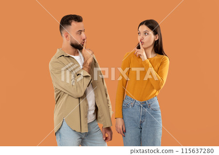 A young man and woman stand facing each other, both holding a finger to their lips, signaling for silence against a solid orange background. They seem to be engaged in a quiet conversation or secret. A young man and woman stand facing each other, both holding a finger to their lips, signaling for silence against a solid orange background. They seem to be engaged in a quiet conversation or secret. 115637280