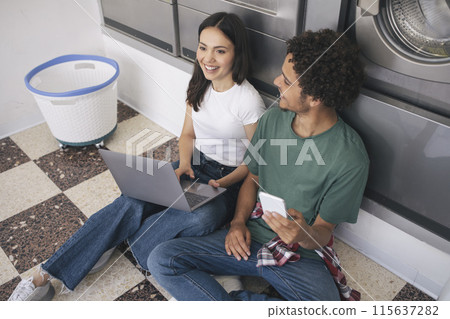 Leisure At Laundry. Cheerful Students Couple With Laptop And Smartphone Sitting In Washing Room With Industrial Washer Machines, Waiting For Clean Clothes At Laundrette Indoors 115637282