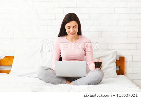 A woman is sitting on a bed, working on a laptop while holding a cup of coffee. She seems focused and engaged in her work, with the morning light filtering through the window. 115637321