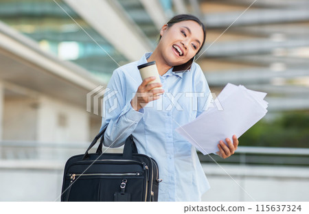 A cheerful Chinese businesswoman, wearing a light blue shirt, holds a cup of coffee and some documents as she walks confidently with a black wheeled briefcase, have phone conversation A cheerful Chinese businesswoman, wearing a light blue shirt, holds a cup of coffee and some documents as she walks confidently with a black wheeled briefcase, have phone conversation 115637324