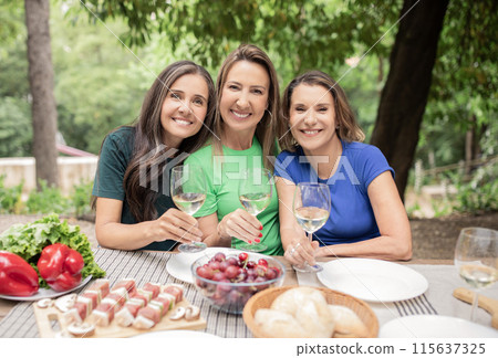 Portrait Of Three Mature Women Enjoying Barbeque Party Outdoors, Happy Female Friends Drinking White Wine And Smiling, Beautiful Ladies Sitting At Table, Holding Glasses And Looking At Camera 115637325