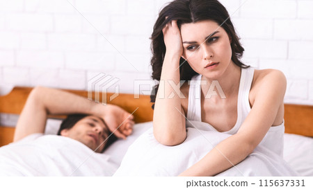A woman sits on a bed, looking worried, with her hand on her head. Her expression is serious and she appears troubled. In the background, a man lies on the bed, asleep. 115637331