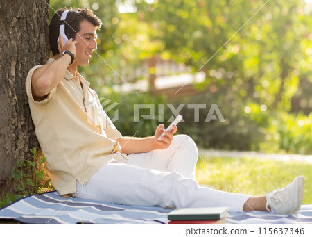 Positive relaxed happy handsome millennial man wearing casual outfit chilling on blanket under tree at park, enjoying sunny summer day, using cell phone and wireless headphones, copy space 115637346
