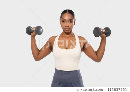 Black woman in a white tank top and gray shorts lifts weights during a workout. She is lifting two grey dumbbells, one in each hand, with her arms extended out to her sides. 115637361