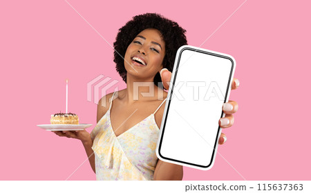 African American young woman with curly hair is holding a piece of birthday cake in one hand and a smartphone with a blank screen in the other hand. The background is a solid pink color. 115637363