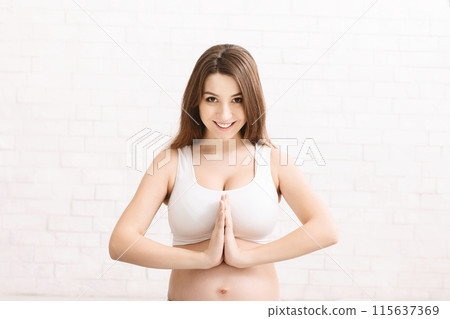 A smiling pregnant woman is performing a yoga pose with hands in a prayer position, set against a bright, sunlit indoor background. She appears calm and relaxed, closeup 115637369