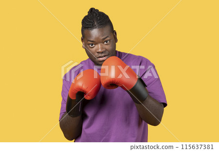 Confident young black boxer man wearing red boxing gloves, ready for combat standing over yellow studio background, looking at camera, portrait shot. Sport training, workout concept Confident young black boxer man wearing red boxing gloves, ready for combat standing over yellow studio background, looking at camera, portrait shot. Sport training, workout concept 115637381