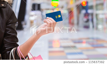 A woman is standing in a busy shopping mall, holding a credit card in one hand. She appears focused on making a purchase or payment, surrounded by shops and other shoppers, cropped A woman is standing in a busy shopping mall, holding a credit card in one hand. She appears focused on making a purchase or payment, surrounded by shops and other shoppers, cropped 115637382