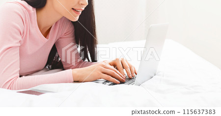 A woman is lying on a bed, focused on using a laptop computer. She is typing and browsing the internet while comfortably positioned. The room is softly lit, cropped 115637383