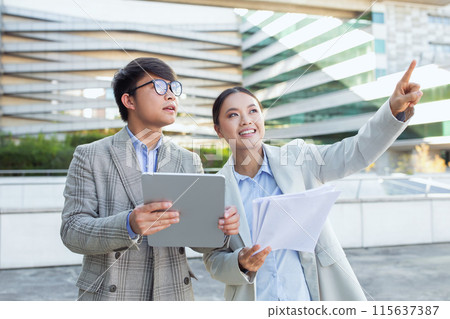 Two young Asian business professionals stand together outside an office building. heir attire and gadgets suggest a work-related interaction during daylight, using digital tablet Two young Asian business professionals stand together outside an office building. heir attire and gadgets suggest a work-related interaction during daylight, using digital tablet 115637387