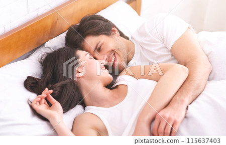 A couple lies in bed, a man and a woman, both smiling and looking at each other. The woman lies on her back with her head resting on the pillow, while the man is on his side, looking down at her 115637403