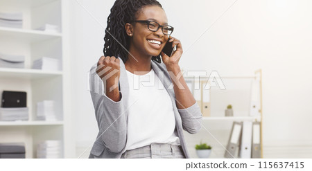 Happy young African American woman in formal attire, a manager standing by the office table, talking on the phone, celebrating success Happy young African American woman in formal attire, a manager standing by the office table, talking on the phone, celebrating success 115637415