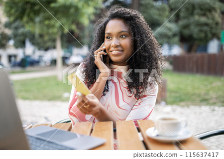 Smiling black woman holding credit card and talking on cellphone outdoors, happy young african american lady sitting at table in cafe, buying online on laptop, enjoying easy payments, closeup Smiling black woman holding credit card and talking on cellphone outdoors, happy young african american lady sitting at table in cafe, buying online on laptop, enjoying easy payments, closeup 115637417