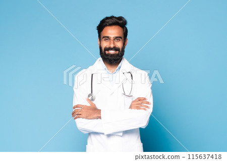 Portrait of friendly indian male doctor in coat with stethoscope on neck posing with folded arms over blue studio background, looking and smiling at camera, free space 115637418
