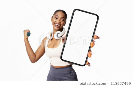 African American young woman with a joyful expression is holding a large smartphone with a blank screen towards the camera. 115637499