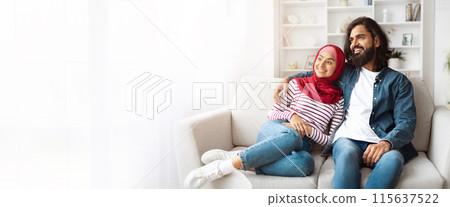A happy Indian couple relaxes on a white sofa in their living room. The woman is wearing a red hijab and a striped shirt while the man is wearing a white t-shirt and a blue denim jacket, copy space A happy Indian couple relaxes on a white sofa in their living room. The woman is wearing a red hijab and a striped shirt while the man is wearing a white t-shirt and a blue denim jacket, copy space 115637522