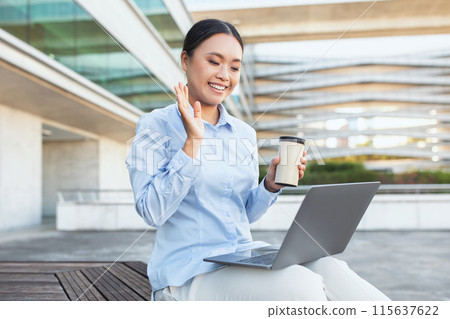 A joyful young Asian woman is seated outside with her laptop on her knees, holding a coffee cup in one hand while waving at the screen during a video call. A joyful young Asian woman is seated outside with her laptop on her knees, holding a coffee cup in one hand while waving at the screen during a video call. 115637622