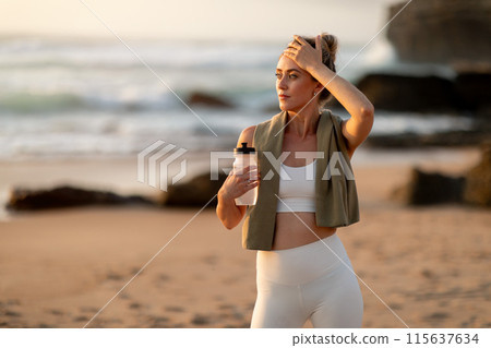 Young tired woman athlete in sportswear holding bottle of water, resting after workout on sea beach, free space. Break, active lifestyle, health care, sport and aqua balance 115637634