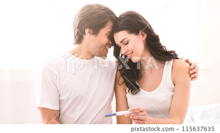 A smiling couple is sitting on a bed at home, with the woman holding a positive pregnancy test. The man has his arm around the woman and they are looking at each other with love and happiness. 115637635