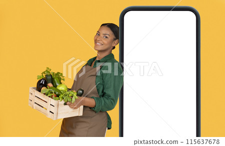 Grocery Market App. Cheerful Black Farmer Woman Leaning On Huge Mobile Phone With Empty Screen, Holding Box Of Fresh Garden Vegetables Over Yellow Studio Background. Mockup, Collage Grocery Market App. Cheerful Black Farmer Woman Leaning On Huge Mobile Phone With Empty Screen, Holding Box Of Fresh Garden Vegetables Over Yellow Studio Background. Mockup, Collage 115637678