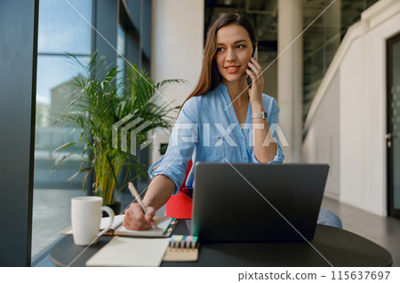 A businesswoman is in her professional workspace, multitasking by taking notes on a call A businesswoman is in her professional workspace, multitasking by taking notes on a call 115637697