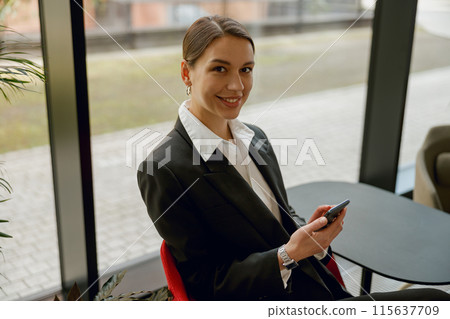 A professional businesswoman is smiling in an office setting while using a smartphone 115637709