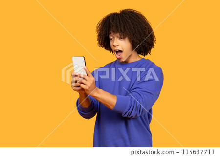 A young man with curly hair and wearing a blue sweatshirt expresses surprise while looking at his smartphone against a bright orange studio background 115637711